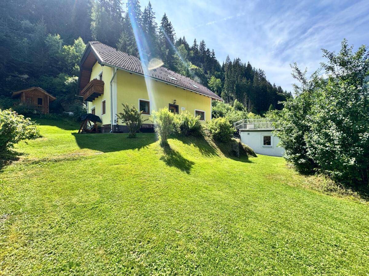 Sonniger Gartenbereich mit weitläufiger Rasenfläche und Blick auf das gelbe Haus und die Berge.