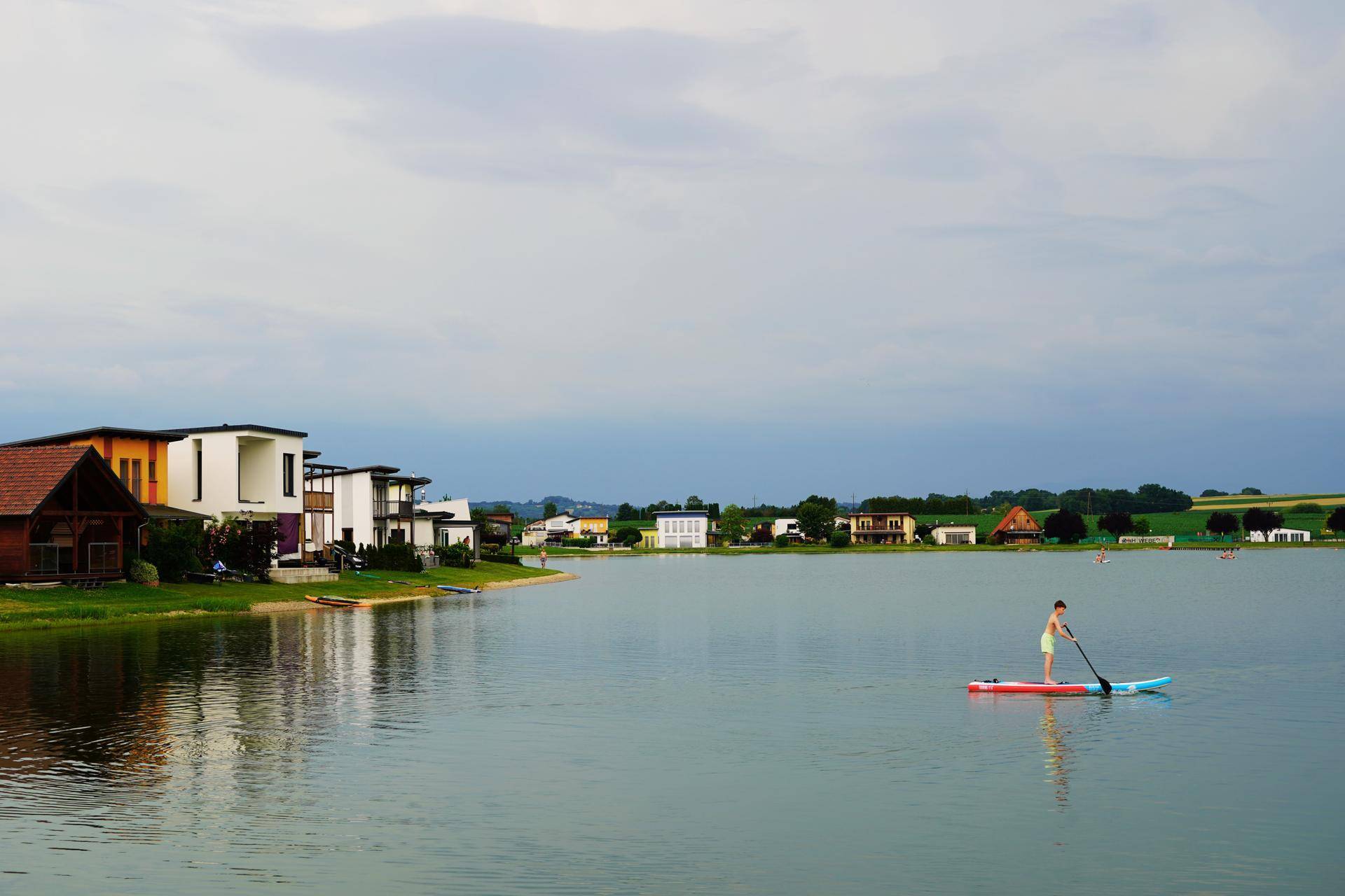 Seeufer mit modernen Häusern und einer Person beim Stand-Up-Paddling an einem bewölkten Tag.