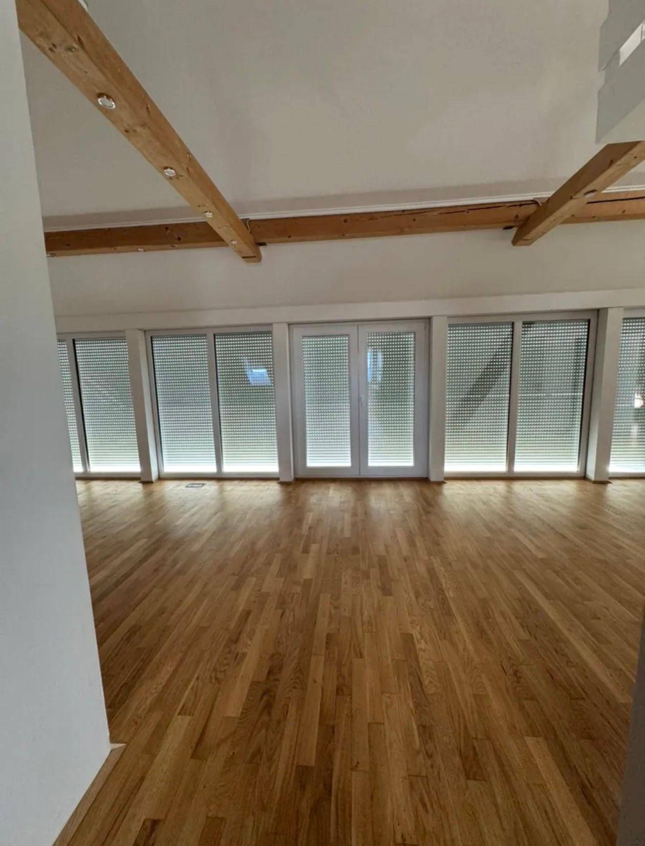 Heller Wohnbereich mit großen Fensterfronten und heruntergelassenen Jalousien, ausgestattet mit Parkettboden.