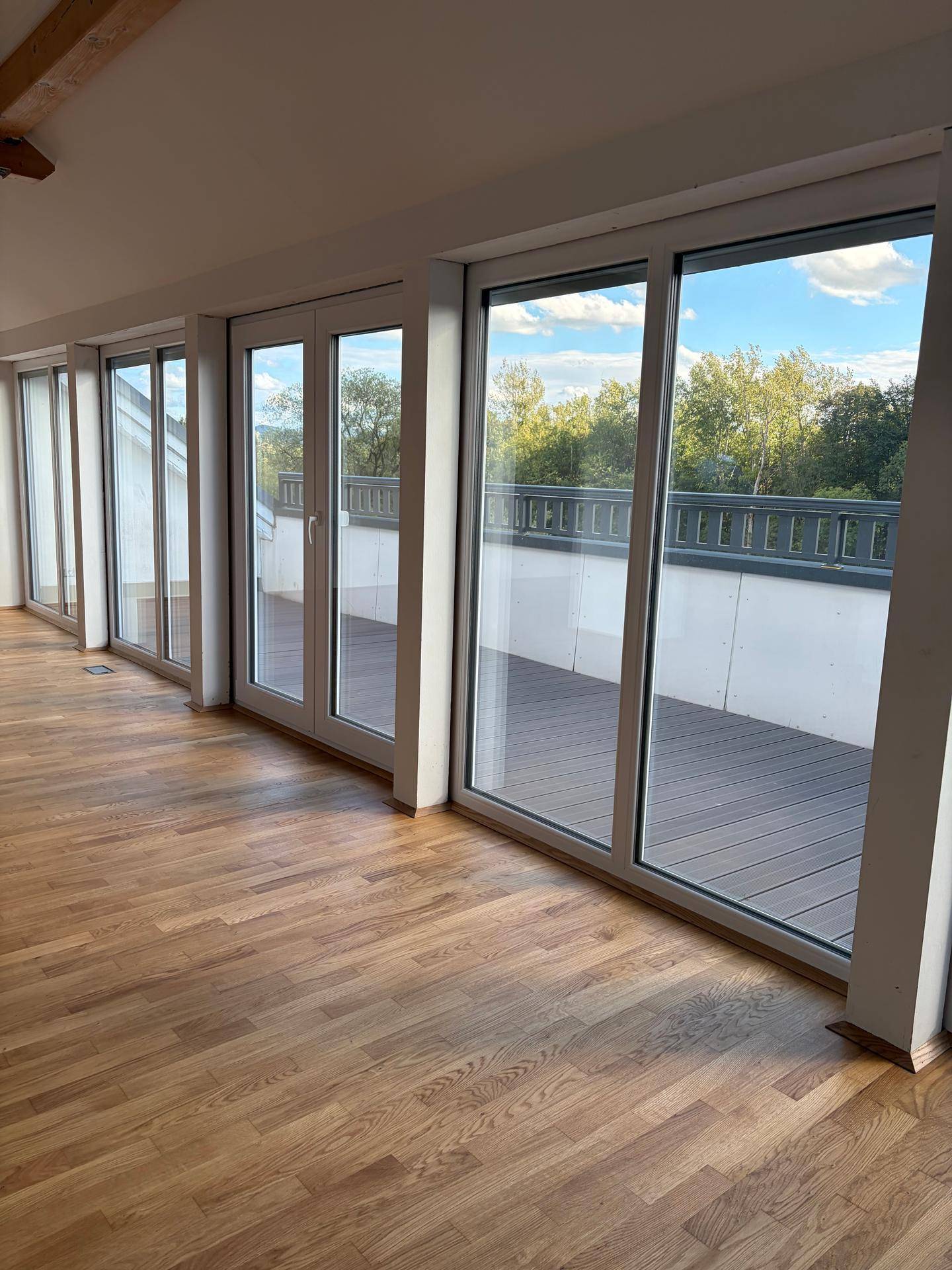 Lichtdurchfluteter Raum mit großen Glasfronten, die Zugang zu einer geräumigen Terrasse und einem grünen Ausblick bieten.
