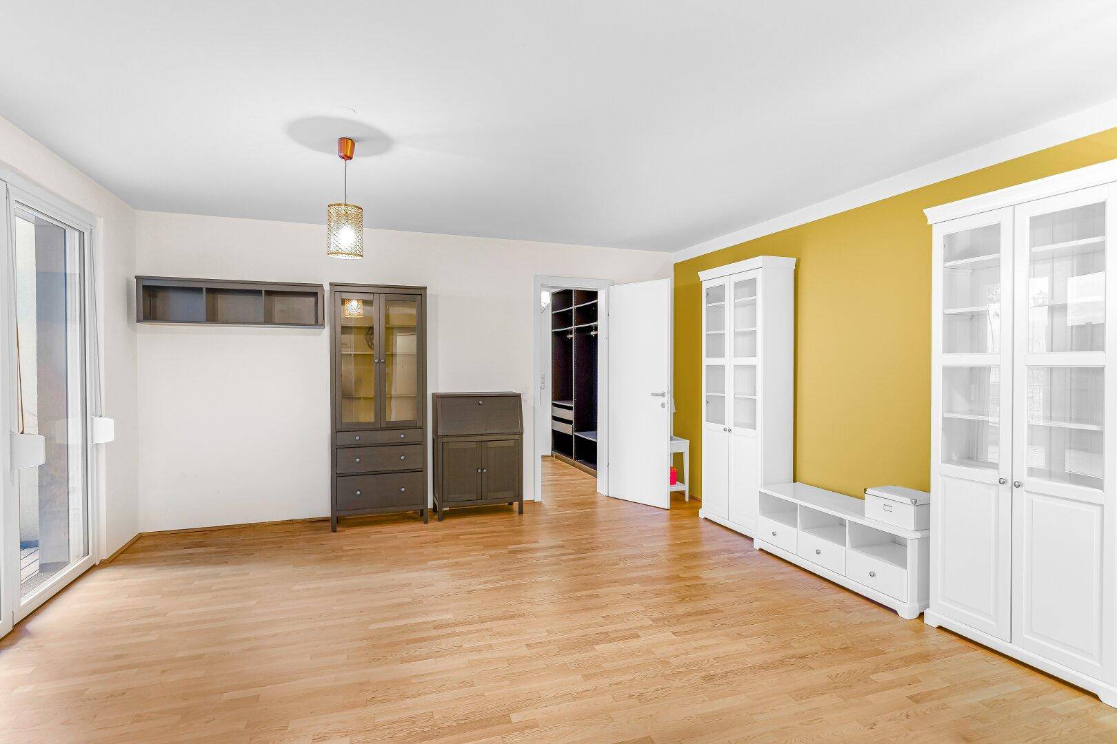Geräumiges Zimmer mit Holzboden, gelber Akzentwand und Zugang zu einem begehbaren Kleiderschrank.