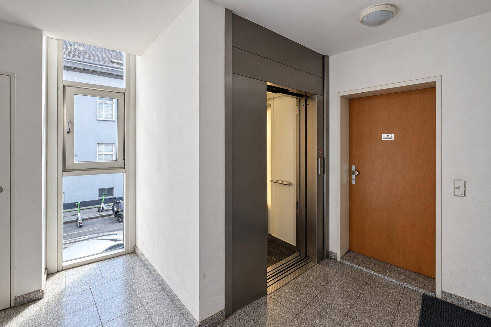 Eingangsbereich mit Aufzug, großem Fenster und gefliestem Boden.