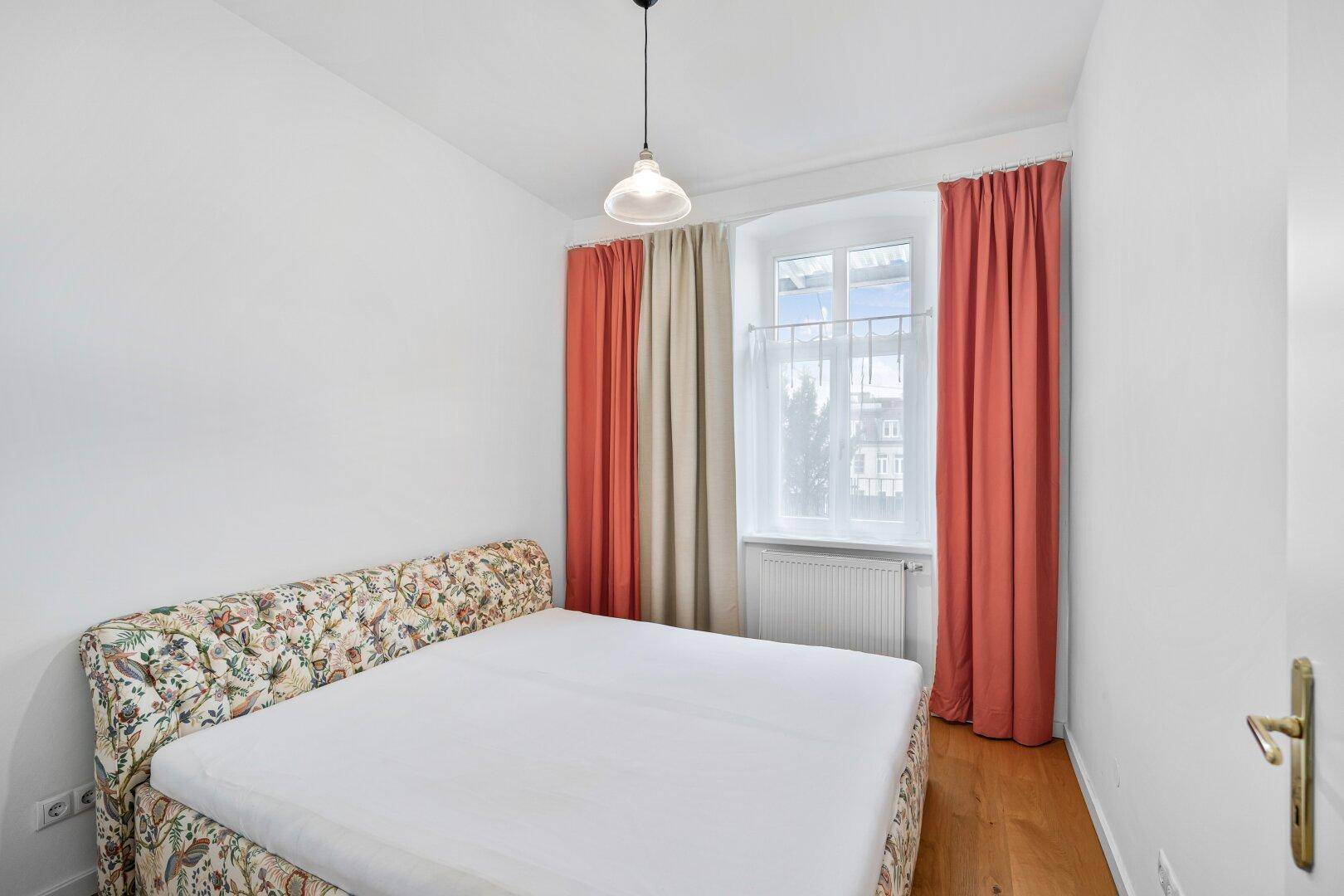 Gemütliches Schlafzimmer mit floral gemustertem Bett, Parkettboden und Fenster.