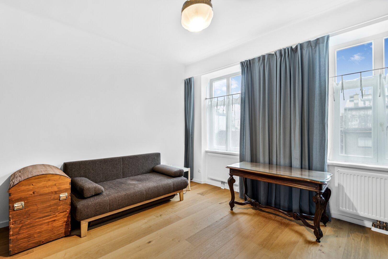 Wohnzimmer mit Sofa, Holztruhe und Schreibtisch, hell durch große Fenster.