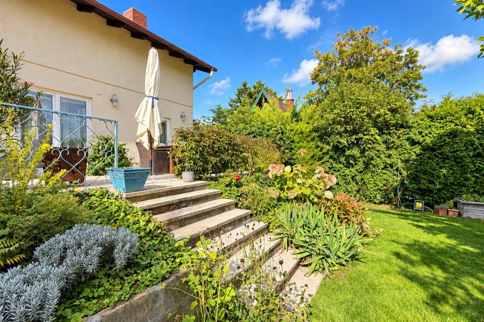 Gepflegter Gartenbereich mit Steintreppe, Blumenbeeten und einer sonnigen Terrasse.