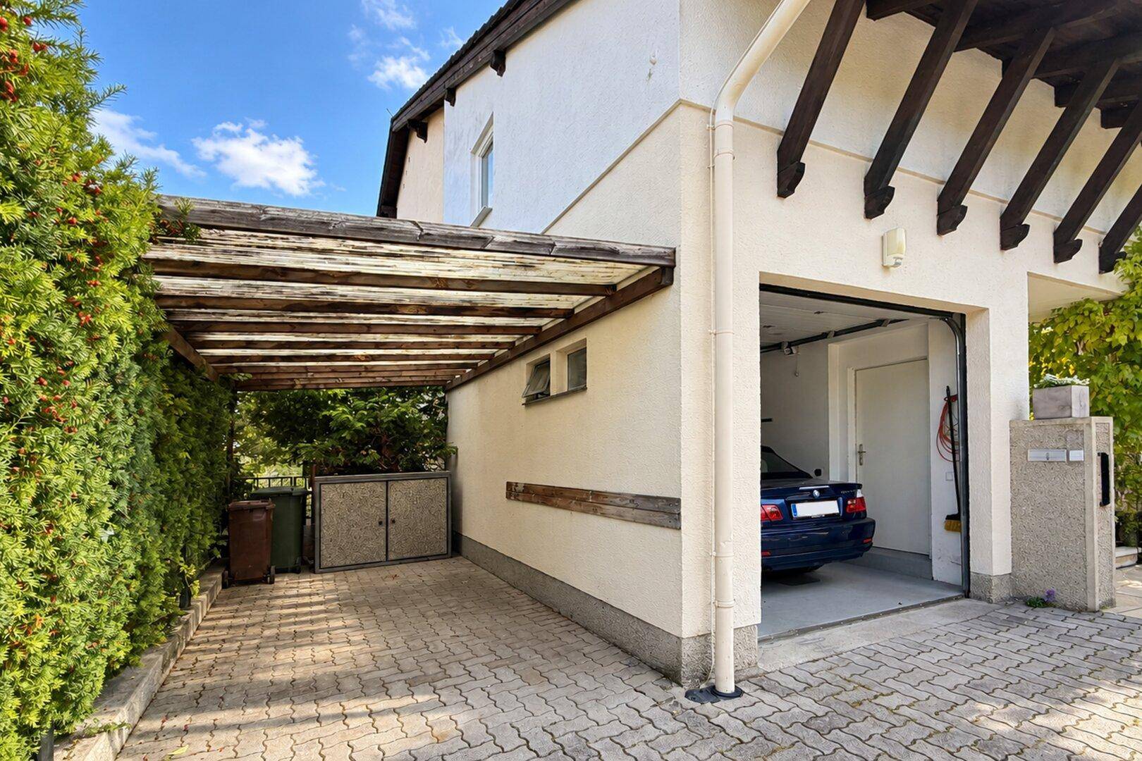 Überdachter Carport und Garage mit Zufahrt, umgeben von einer hohen Hecke.