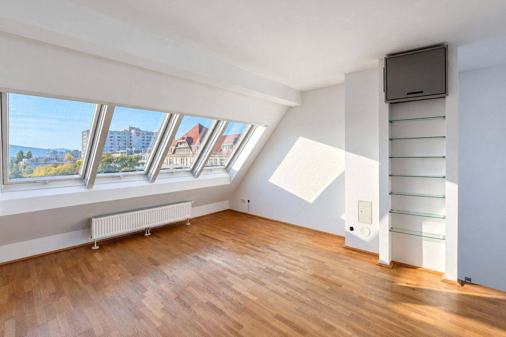Geräumiger, heller Raum mit Holzboden und großen Dachfenstern, die einen weiten Blick über die Stadt bieten.
