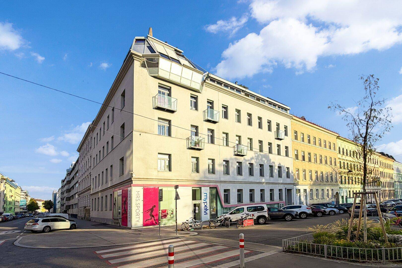Mehrstöckiges Gebäude mit Geschäften im Erdgeschoss, Balkonen und einem Mansarddach in städtischer Umgebung.
