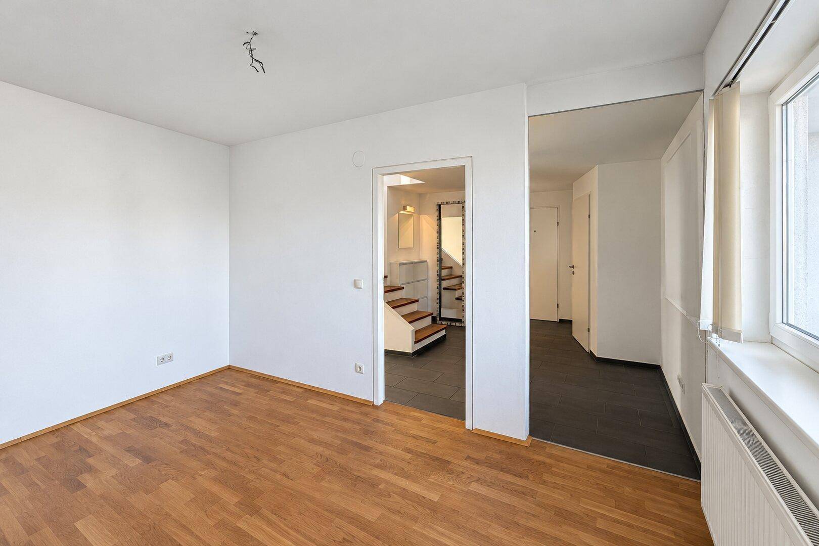 Helles Zimmer mit Holzboden und Fenster, das einen Blick in den Flur mit Treppe bietet.
