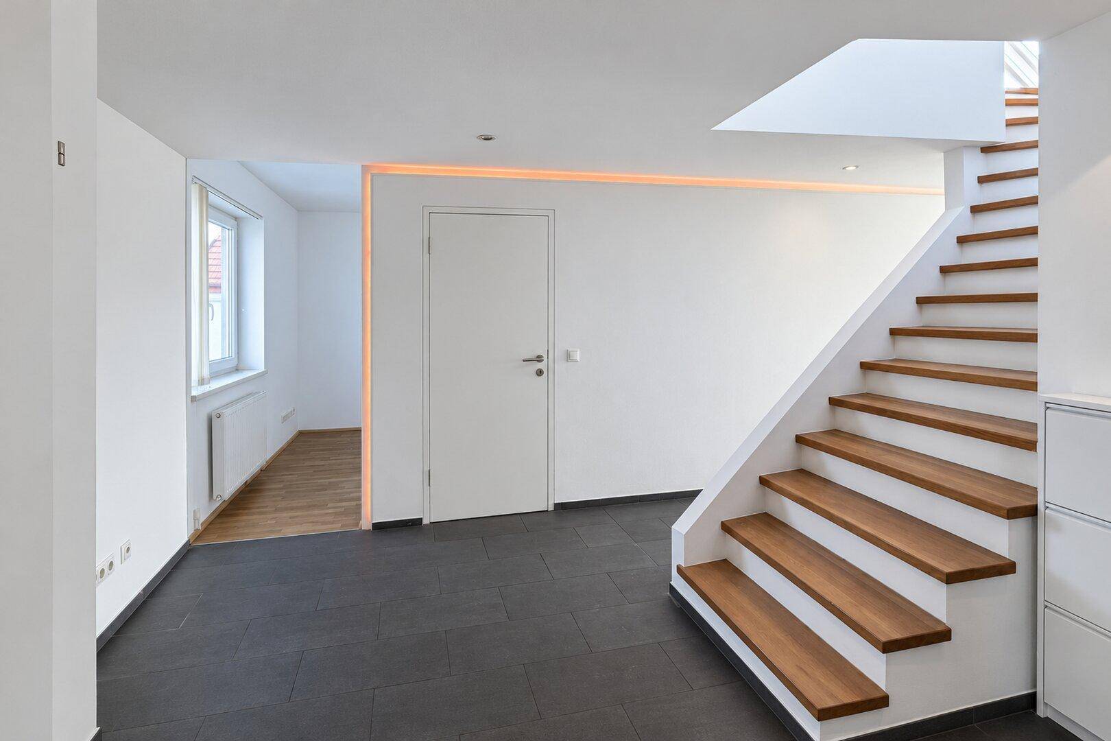 Heller Flur mit dunklem Fliesenboden und einer modernen Holztreppe, beleuchtet durch indirektes Licht.