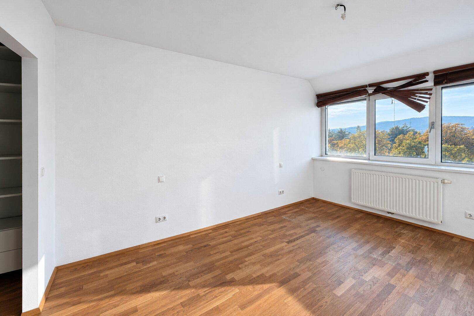 Geräumiges Schlafzimmer mit Holzboden, großen Fenstern und einem angenehmen Blick ins Grüne.
