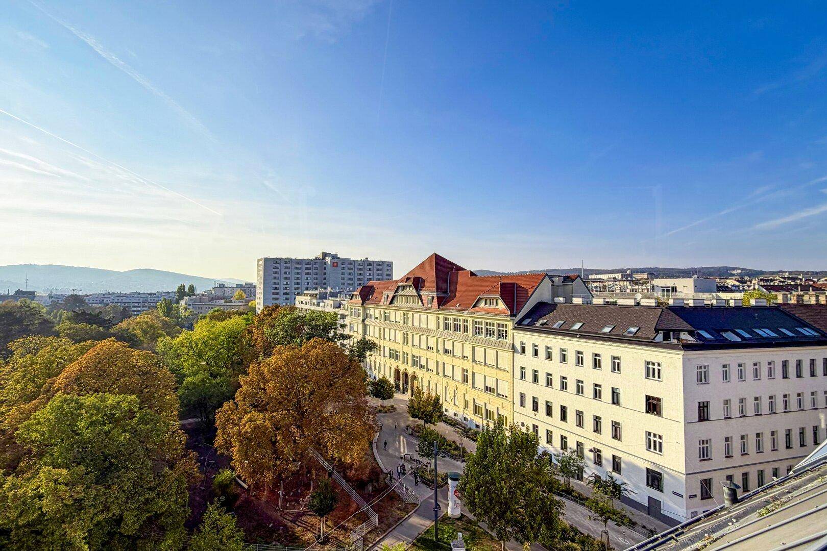 Panoramablick über die Stadt mit grünen Bäumen, Gebäuden und fernen Bergen unter blauem Himmel.