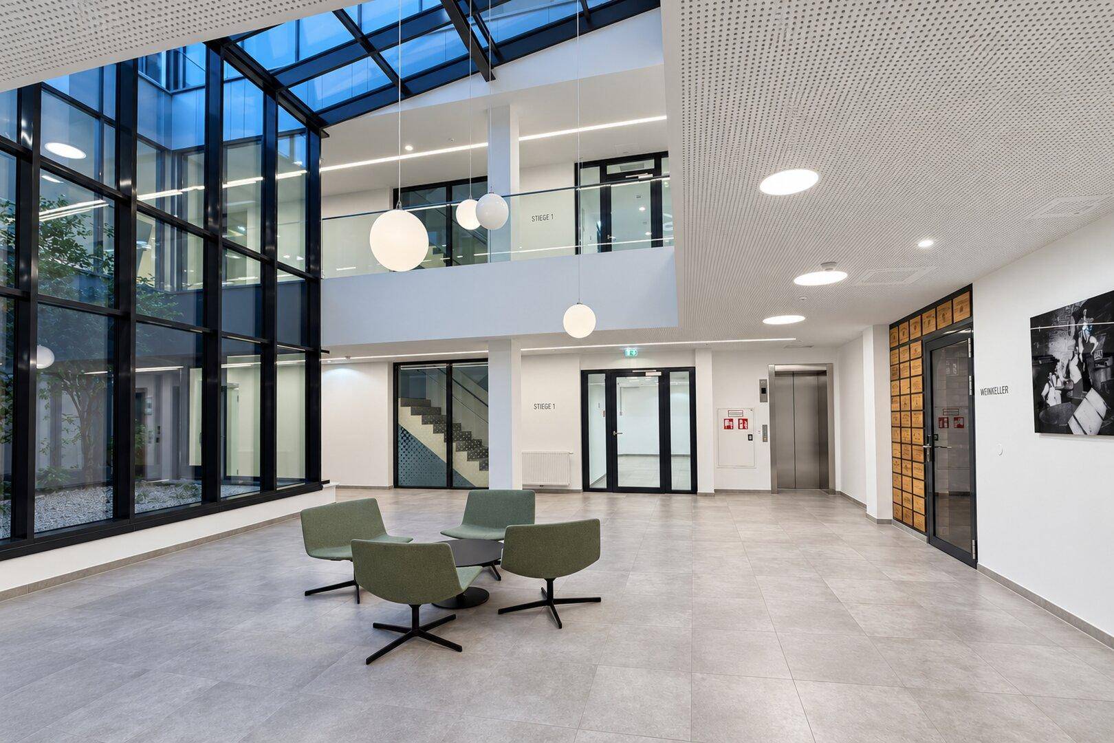Großzügige Lobby mit Glasdach, Sitzgelegenheiten und Aufzug in einem modernen Gebäude.