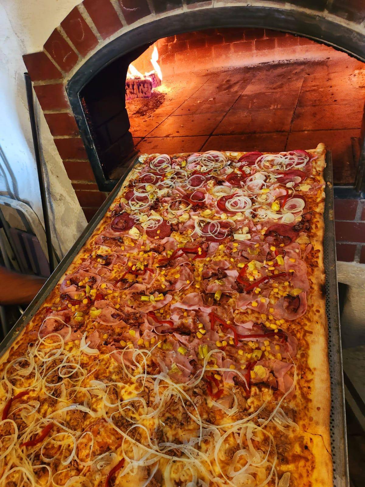 Eine riesige Pizza wird in einen traditionellen, gemauerten Holzofen geschoben, mit loderndem Feuer im Hintergrund.