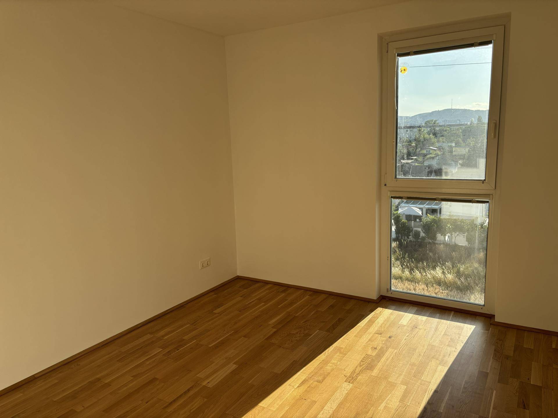 Leeres Zimmer mit Holzparkettboden und großem Fenster mit Blick ins Grüne.