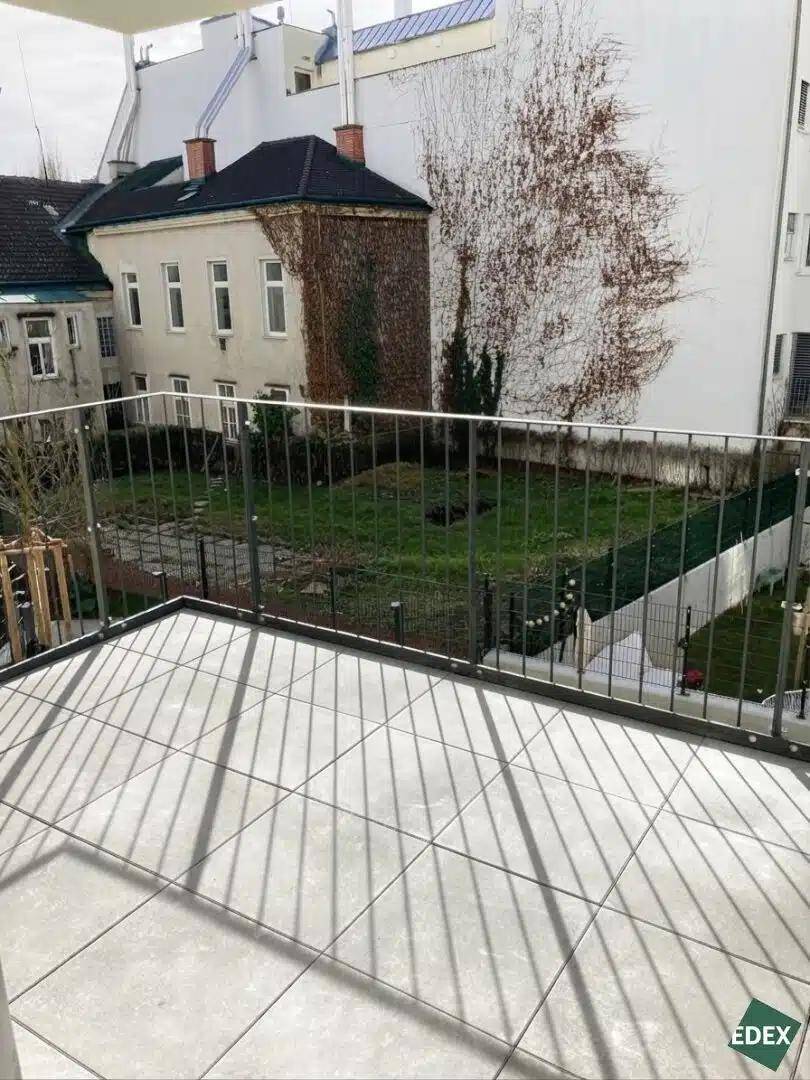 Balkon mit Fliesenboden und Blick auf einen begrünten Innenhof und umliegende Gebäude.