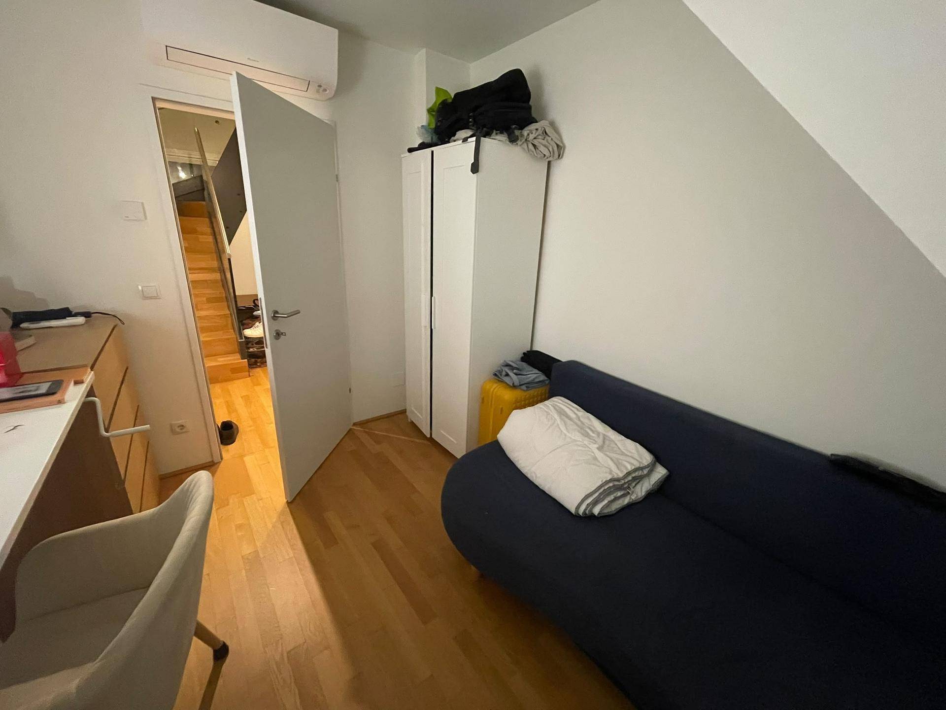 Gemütliches Dachzimmer mit Holzboden, Sofa und Klimaanlage, mit Zugang zum Treppenhaus.