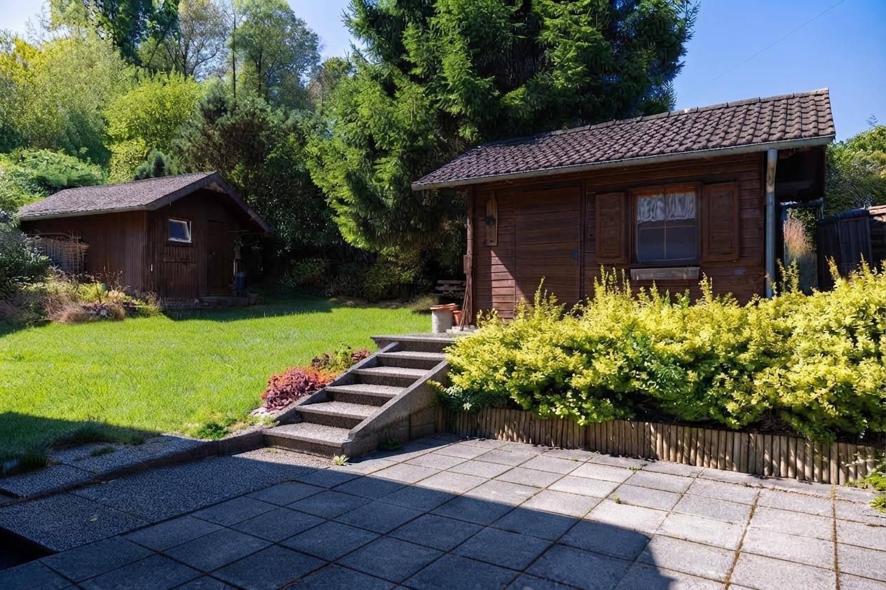 Großer, sonniger Garten mit Rasenfläche, Sträuchern und zwei rustikalen Gartenhäusern aus Holz.