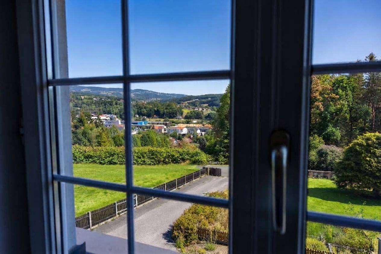 Blick aus einem Fenster mit Sprossen auf eine weite grüne Landschaft und entfernte Häuser.