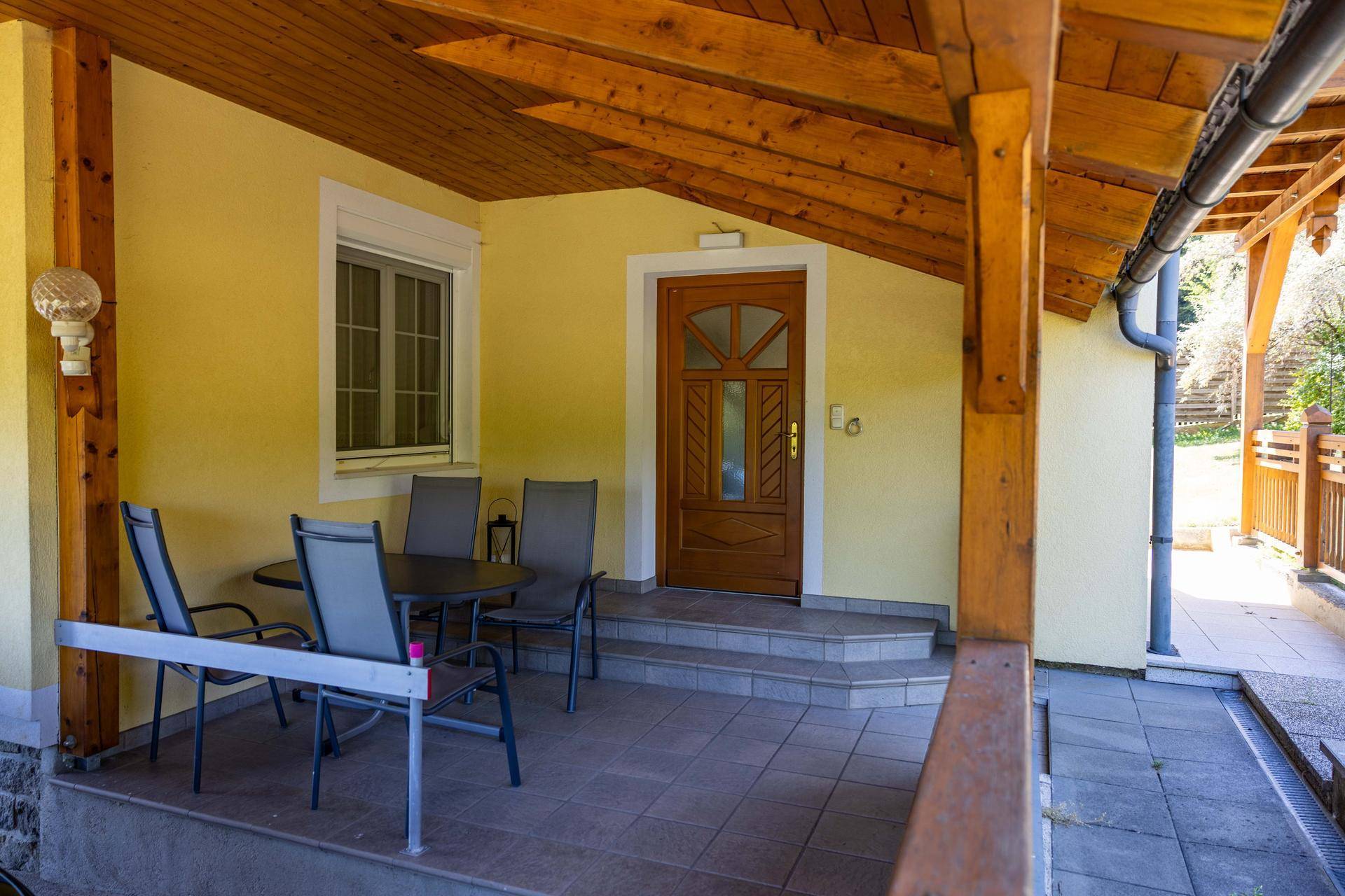 Gemütliche überdachte Terrasse mit Sitzgelegenheiten, Holzbalken und Fliesenboden am Eingangsbereich.