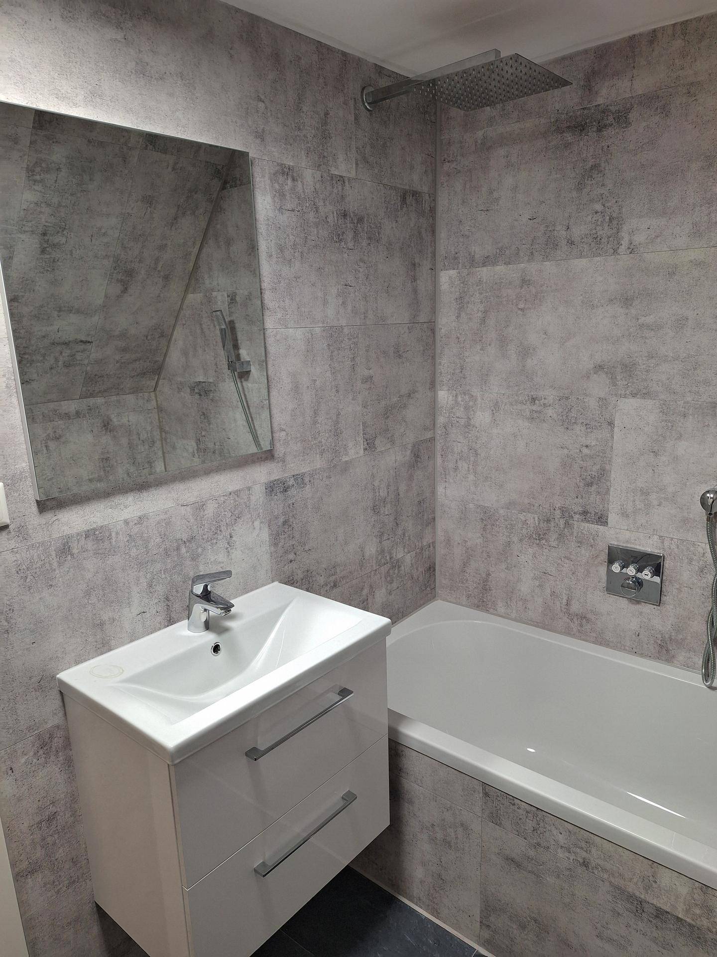 Badezimmer mit Waschtisch, Spiegel, Badewanne und Regendusche, komplett gefliest in Grautönen und funktional.