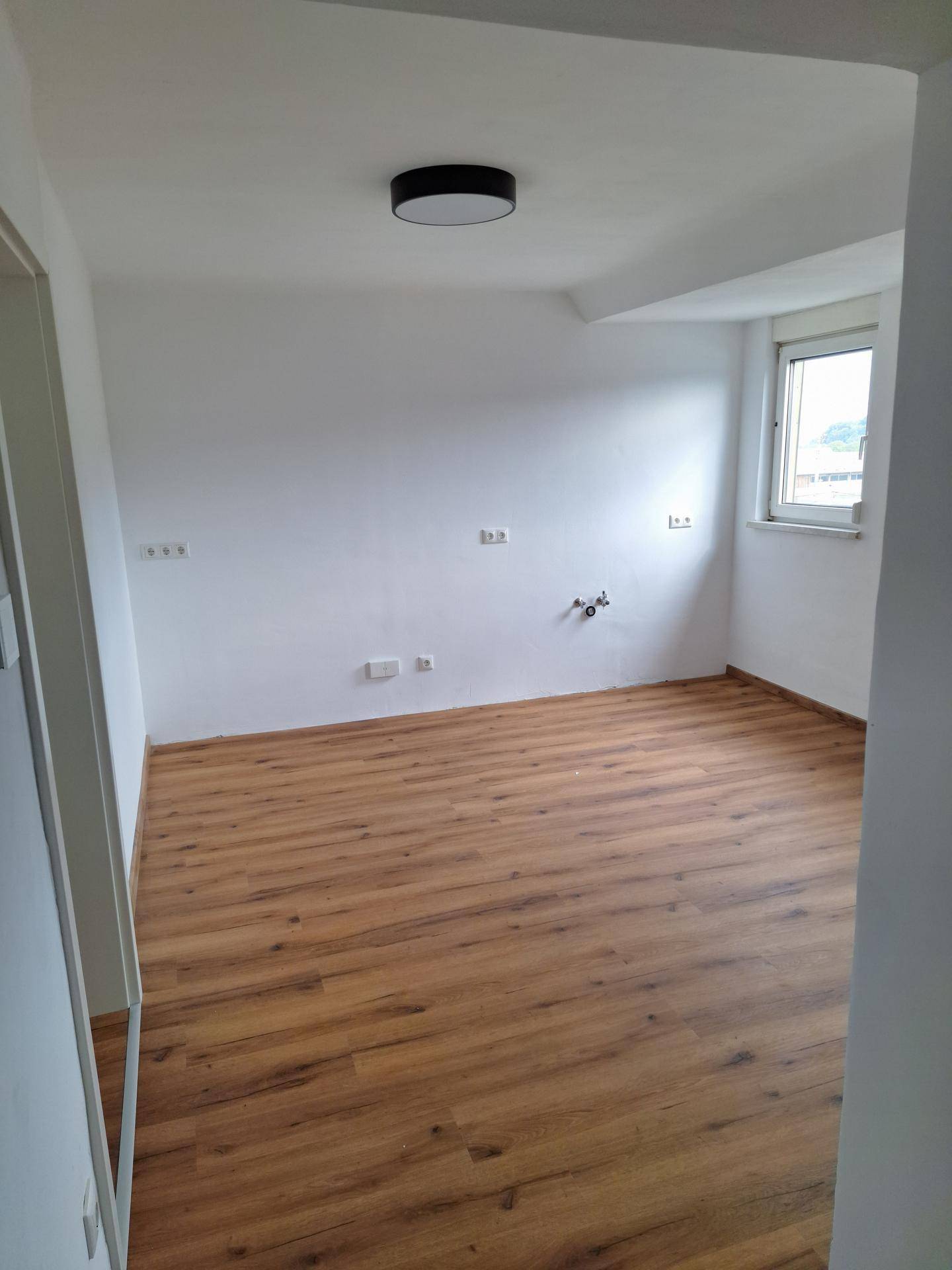 Leerer Raum mit Laminatboden und Fenster, vorbereitet für die Installation einer Küche mit allen Anschlüssen.