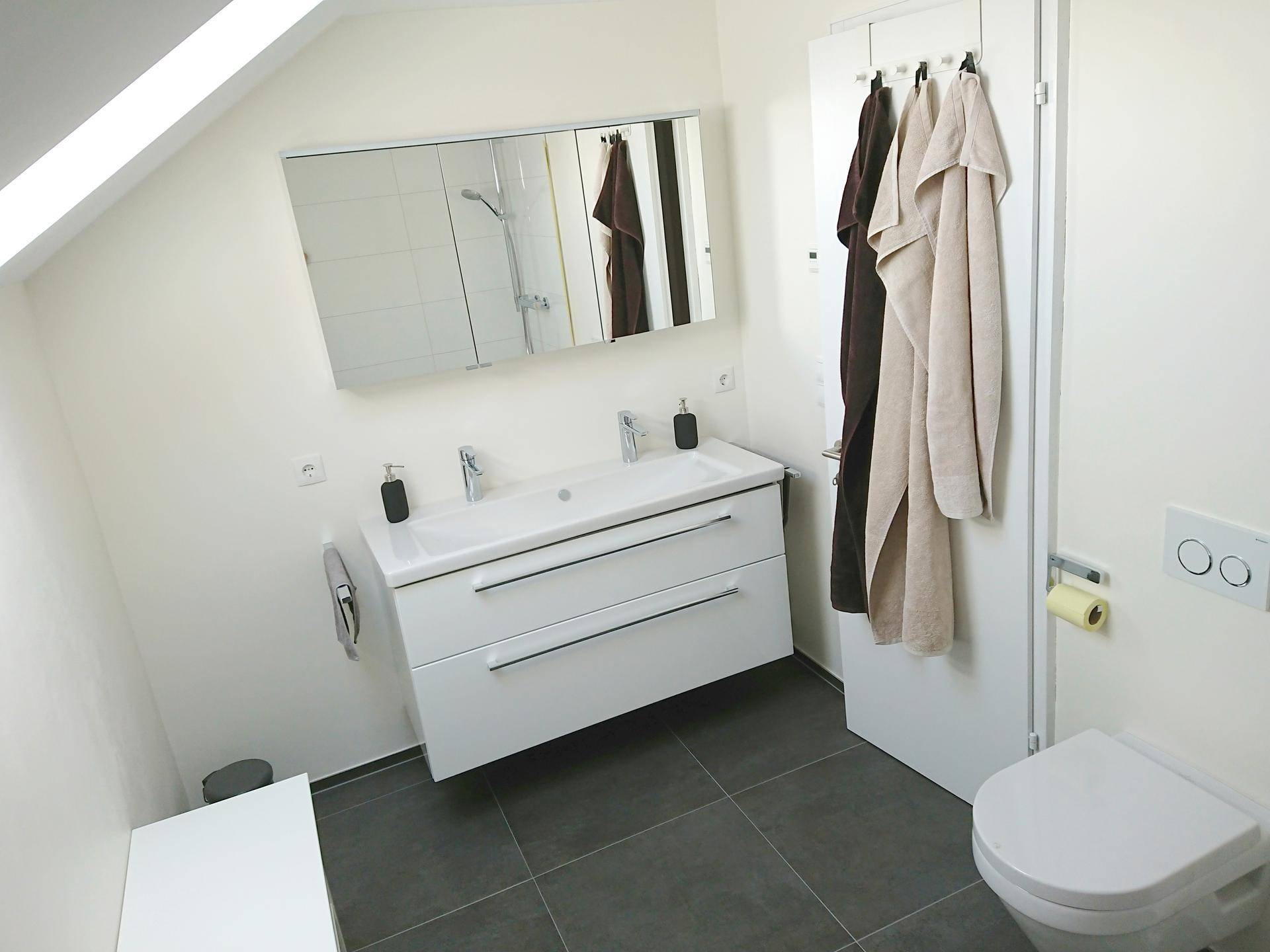 Modernes Badezimmer im Dachgeschoss mit Doppelwaschbecken, Spiegelschrank und dunklen Bodenfliesen.