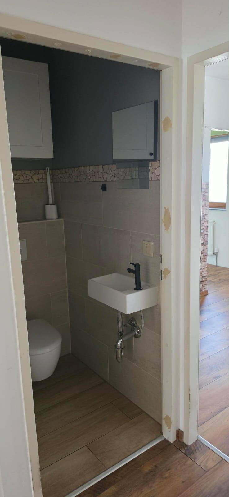 Modernes Gäste-WC mit Wand-WC, kleinem Waschbecken und hellen Fliesen, zugänglich vom Flur.