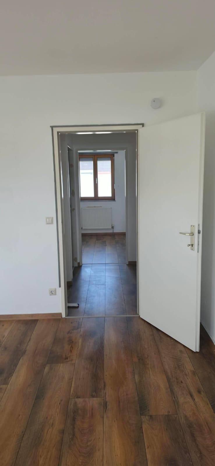 Offene Tür führt von einem Flur in einen weiteren Raum mit Laminatboden und Fenster.