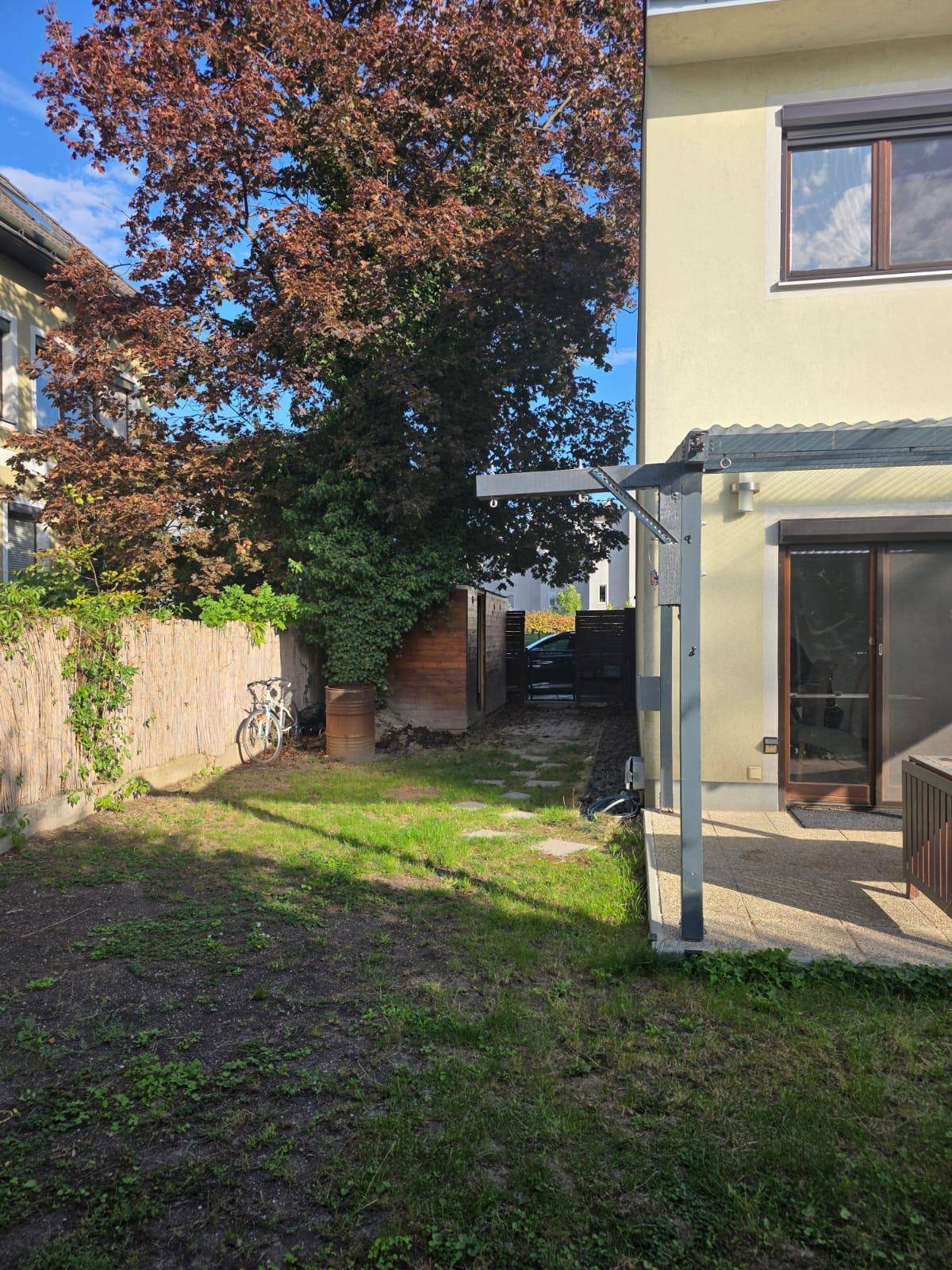 Gartenansicht mit Rasenfläche, überdachter Terrasse und einem großen Baum, der Schatten spendet.