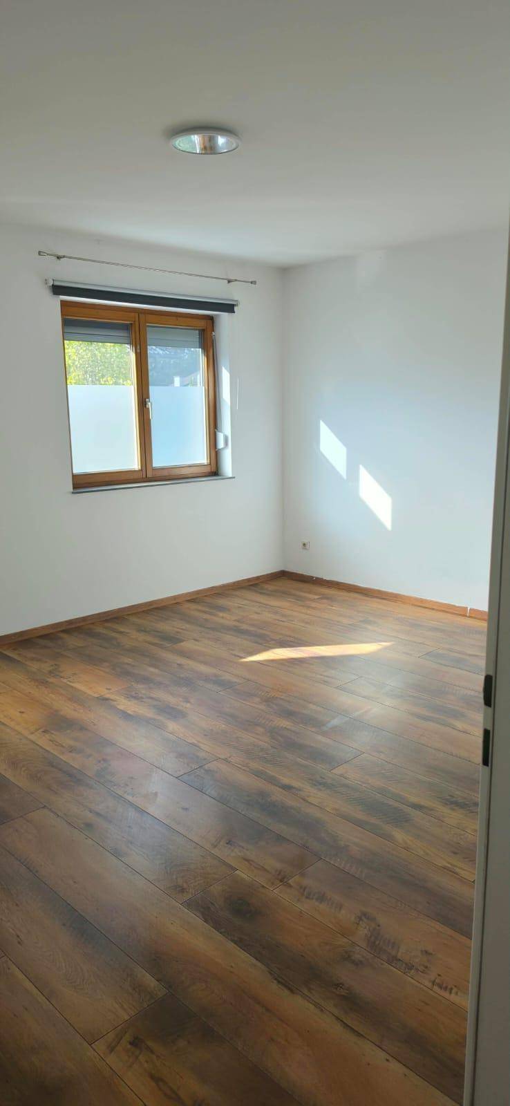 Geräumiger, heller Raum mit Laminatboden und einem Fenster mit Holzrahmen, das viel Tageslicht hereinlässt.