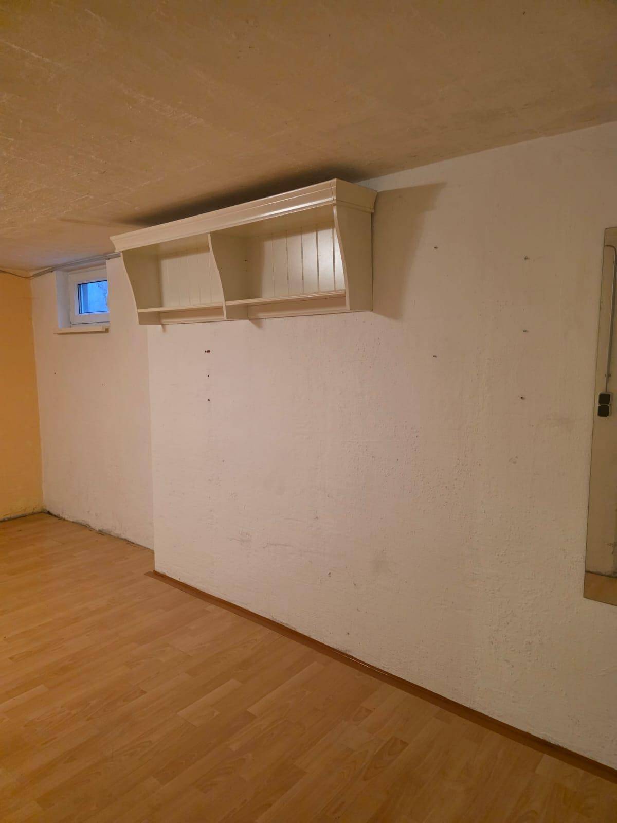 Kellerraum mit hellem Laminatboden, weißen Wänden und einem kleinen Fenster.