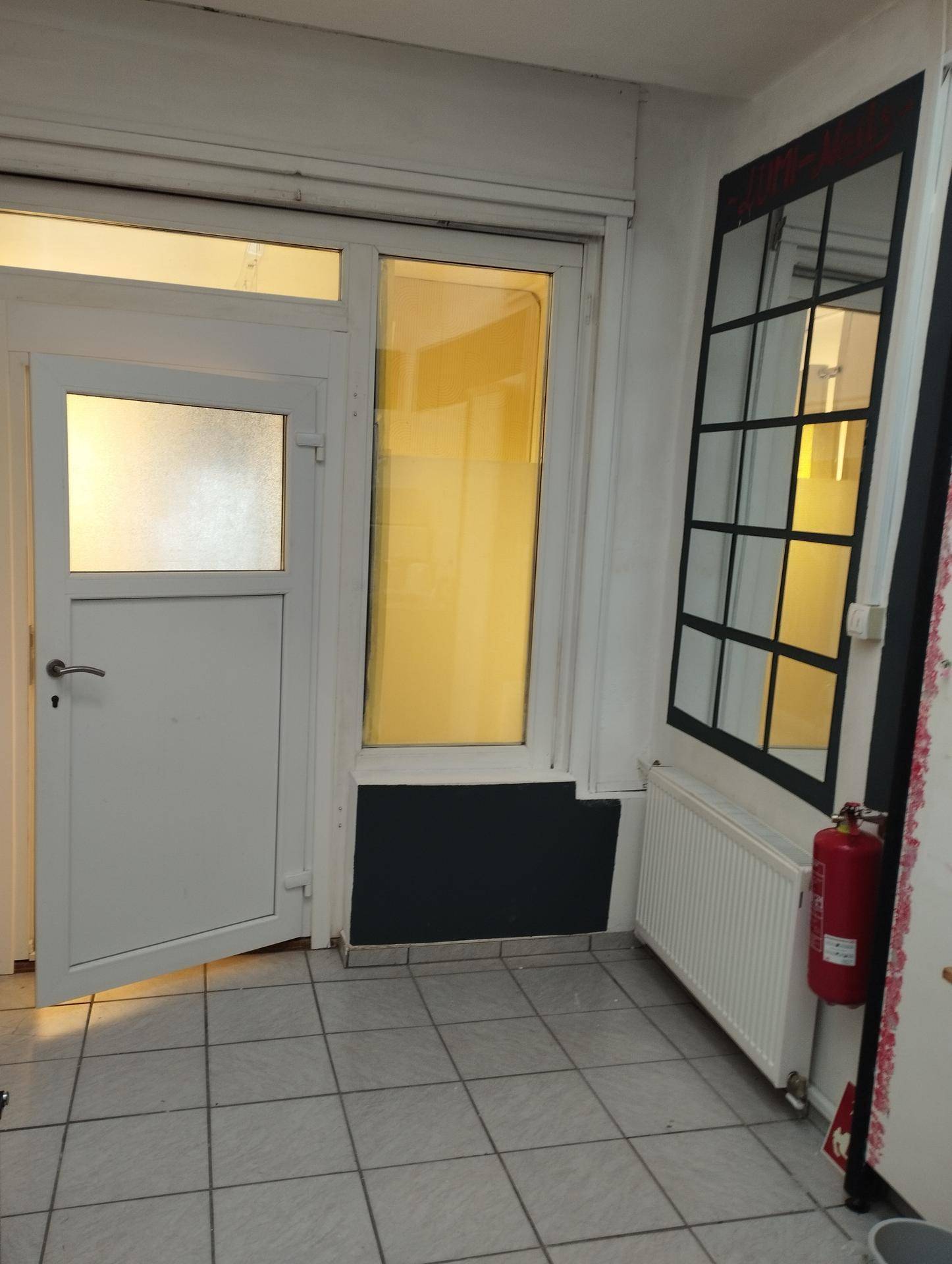 Eingangsbereich mit weißer Tür, hohem Fenster, Heizkörper und Fliesenboden, ausgestattet mit einem Feuerlöscher.