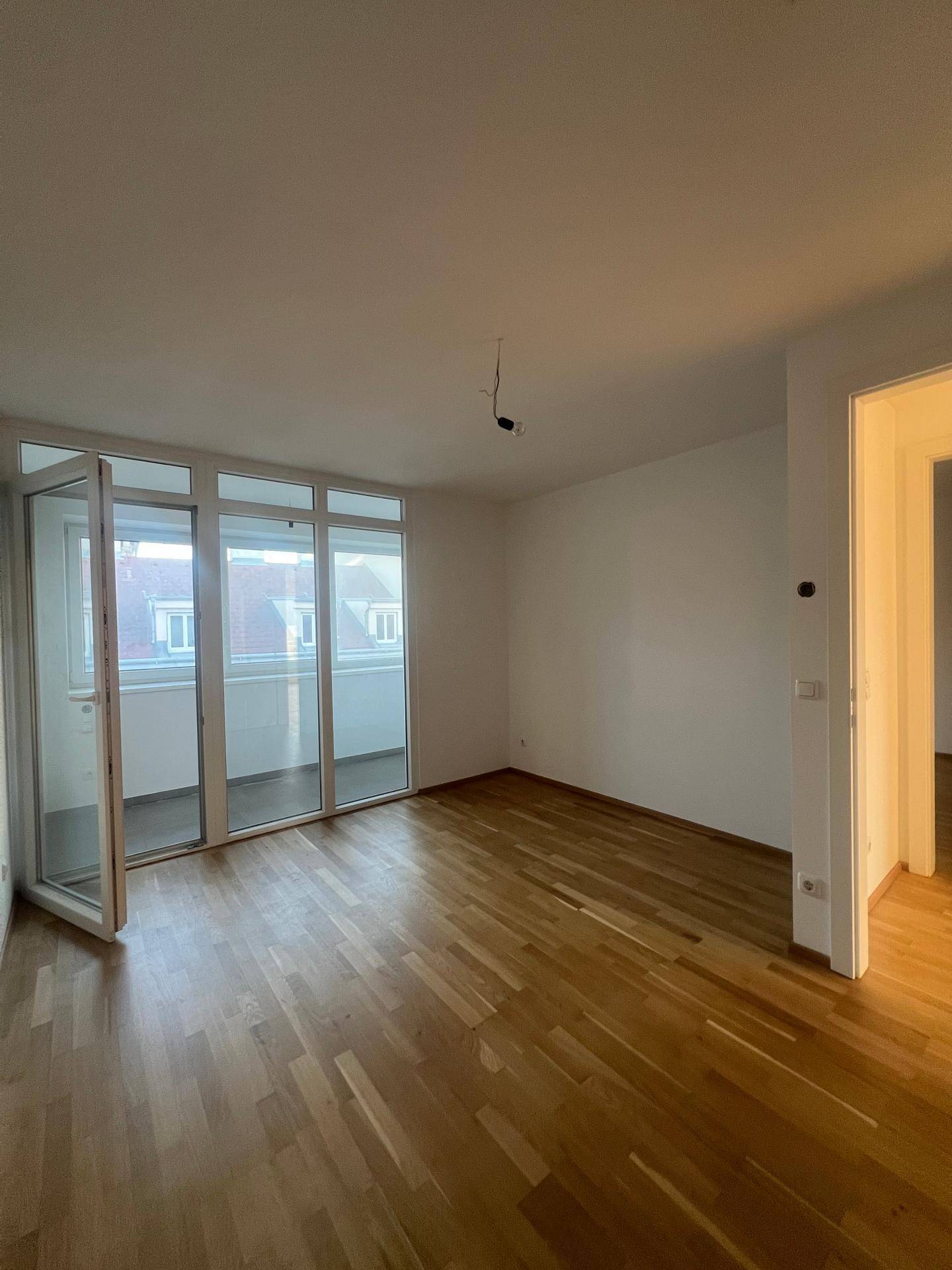 Geräumiges Zimmer mit Holzboden und breiter Fensterfront, die viel Licht spendet und Zugang zum Balkon bietet.