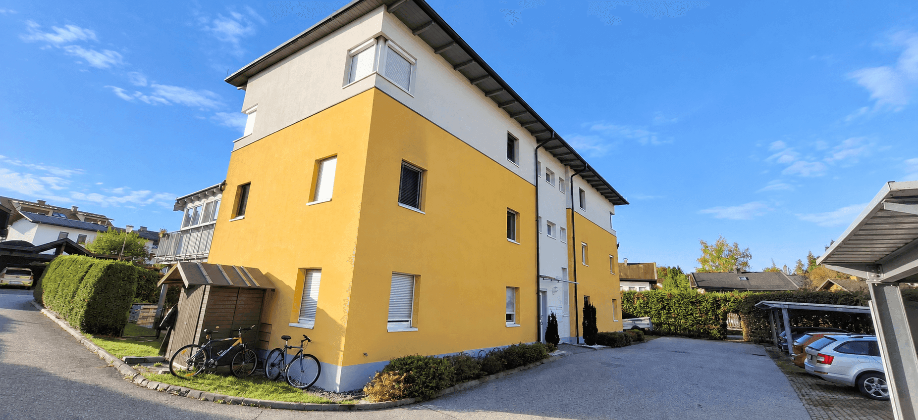 Ansicht des Mehrfamilienhauses mit gelber Fassade, Parkplätzen und Carports im Außenbereich.