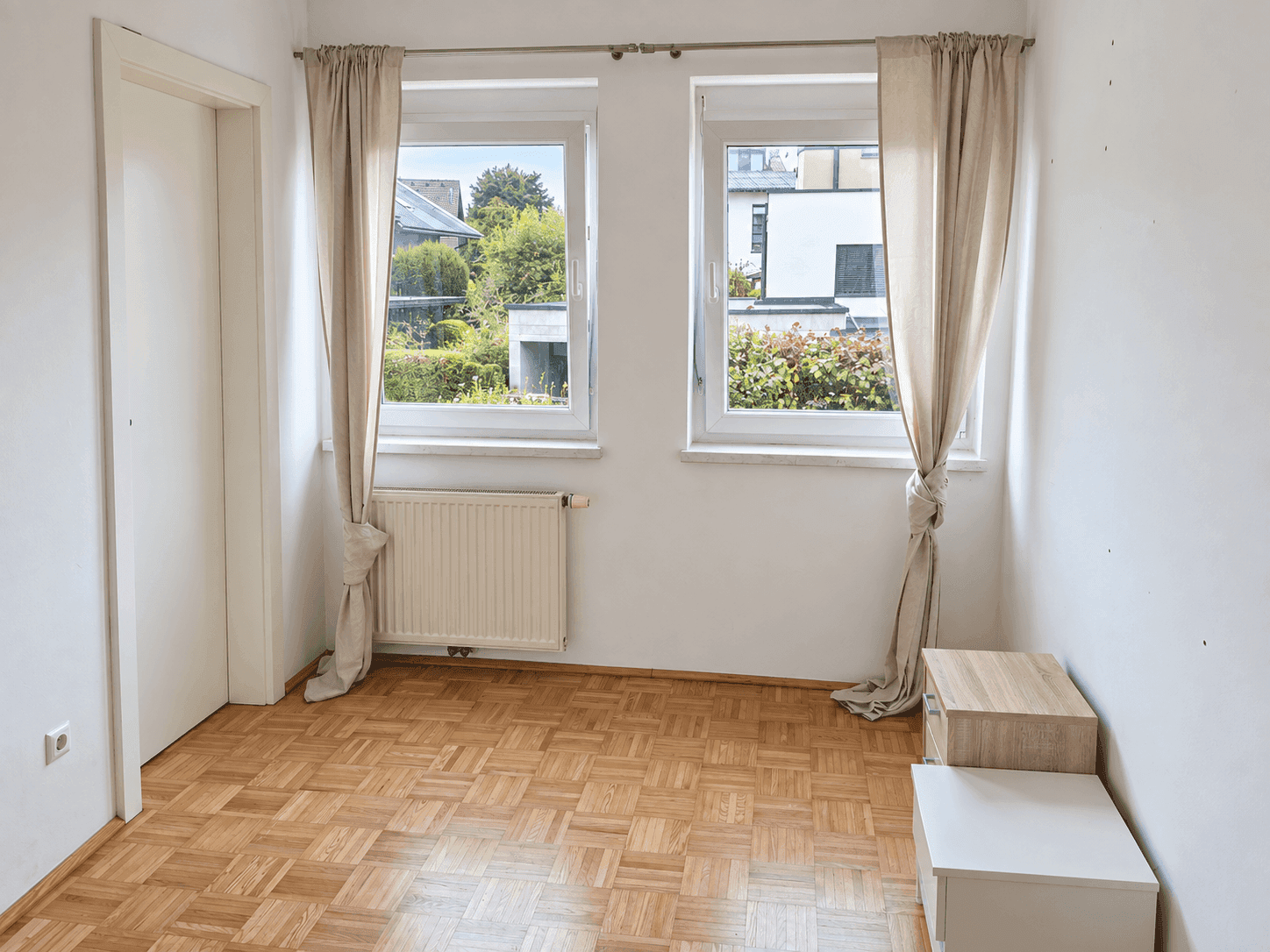 Helles Zimmer mit Parkettboden, zwei Fenstern und Blick auf die umliegende Wohngegend.