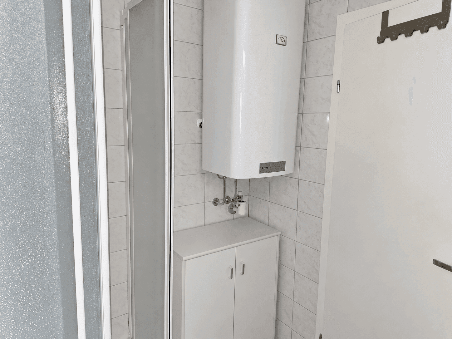 Badezimmer mit Duschkabine und Warmwasserboiler, ausgestattet mit hellen Wandfliesen.