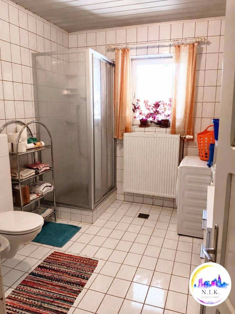 Badezimmer mit weiß gefliesten Wänden, Duschkabine, WC und einem Fenster für Tageslicht.