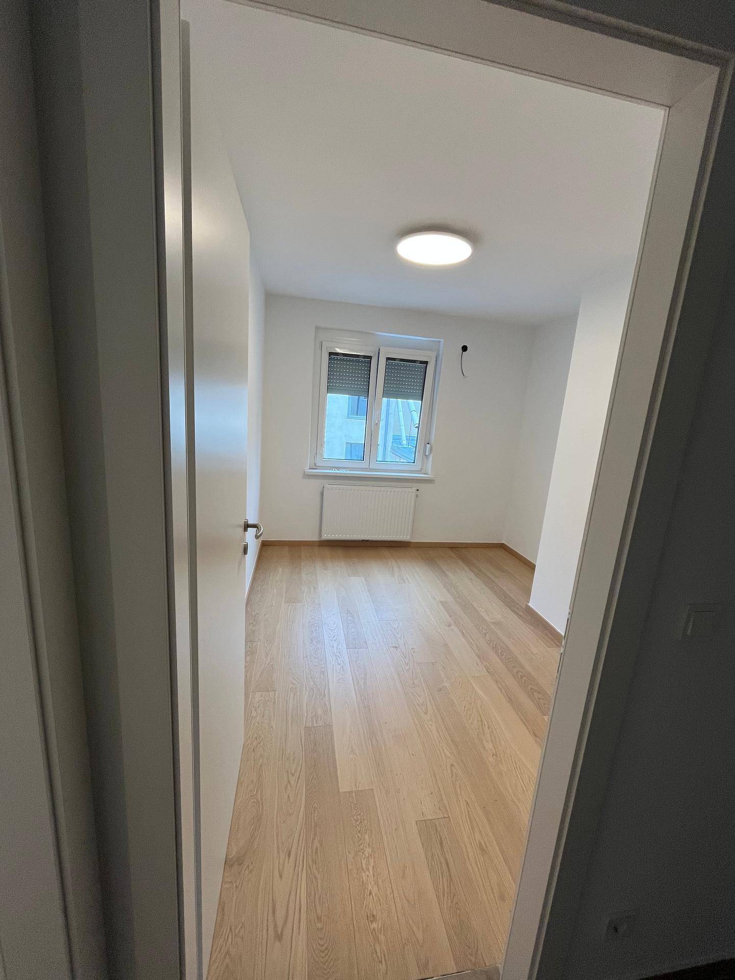 Blick durch eine offene Tür in ein helles Zimmer mit Parkettboden und Fenster.