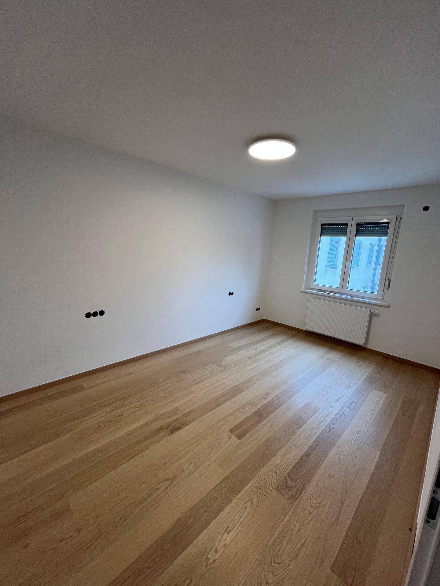 Geräumiges Zimmer mit hellem Parkettboden und Fenster, das viel Tageslicht hereinlässt.