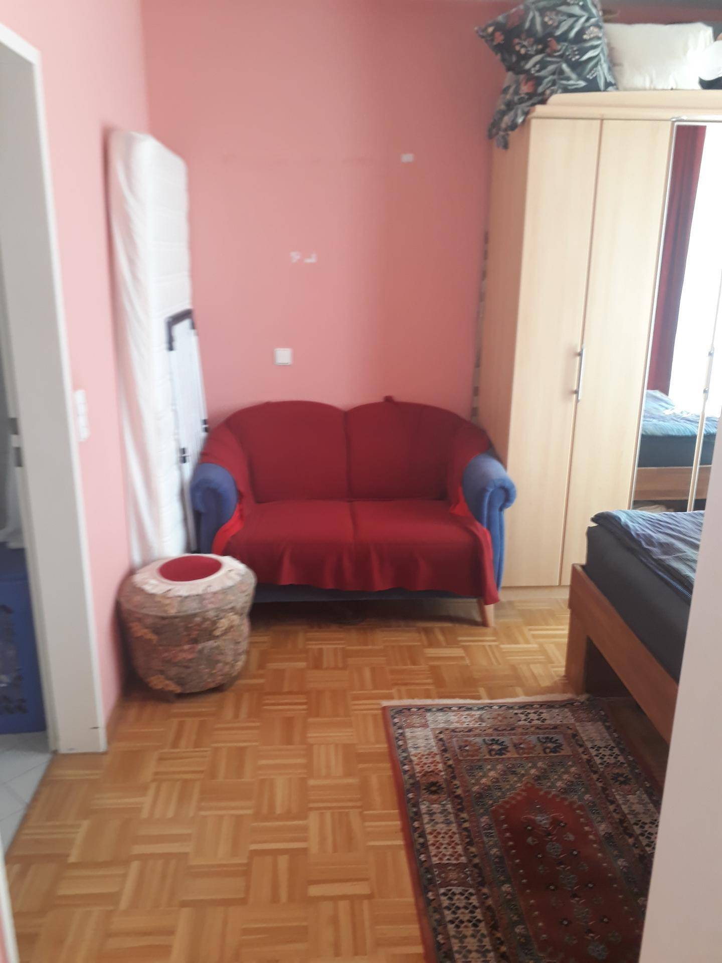 Schlafzimmer mit rotem Sofa, Kleiderschrank und Parkettboden in warmen Farbtönen.