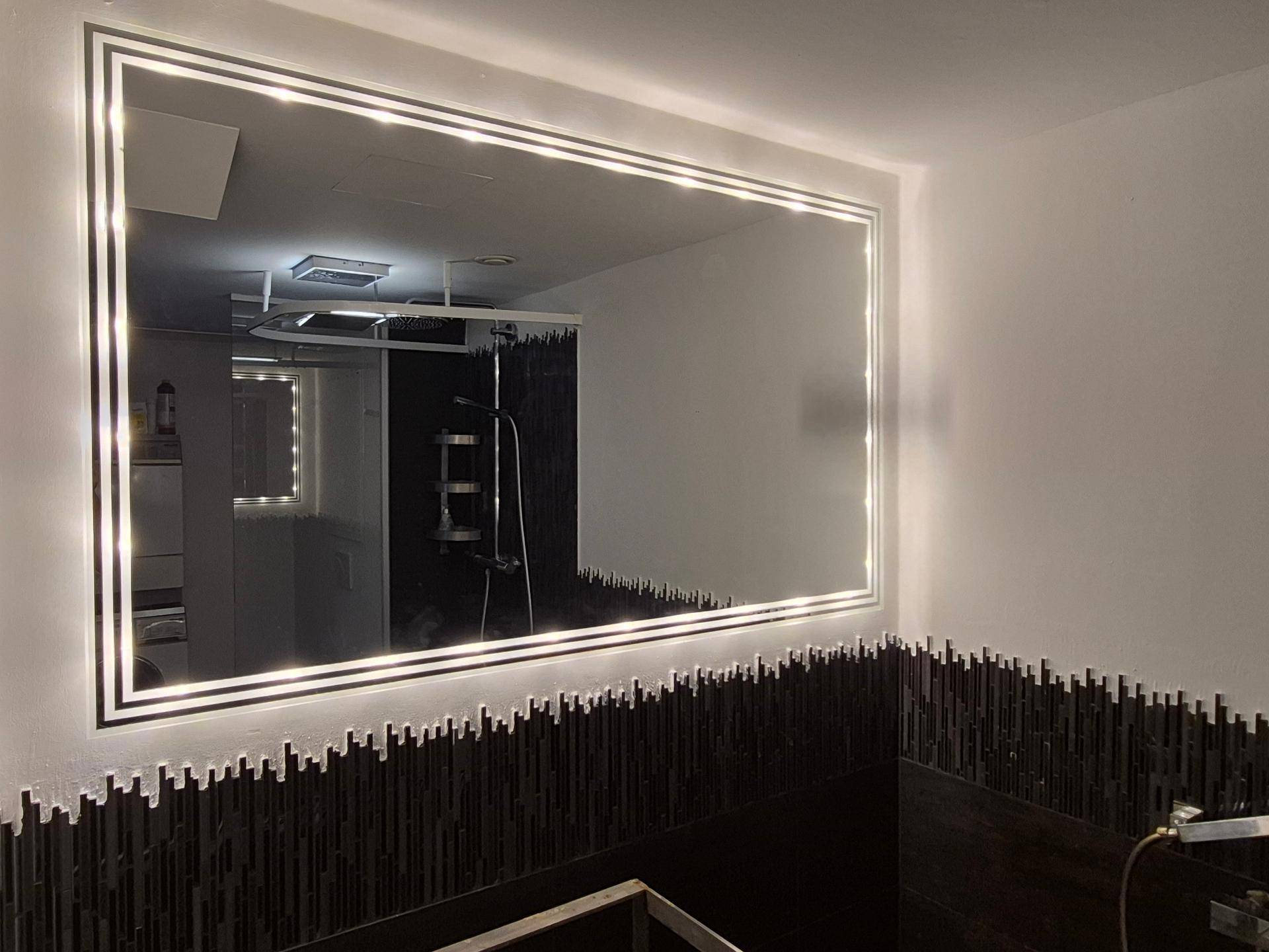 Badezimmer mit großem, beleuchtetem Spiegel und dunklen Mosaikfliesen an der Wand.