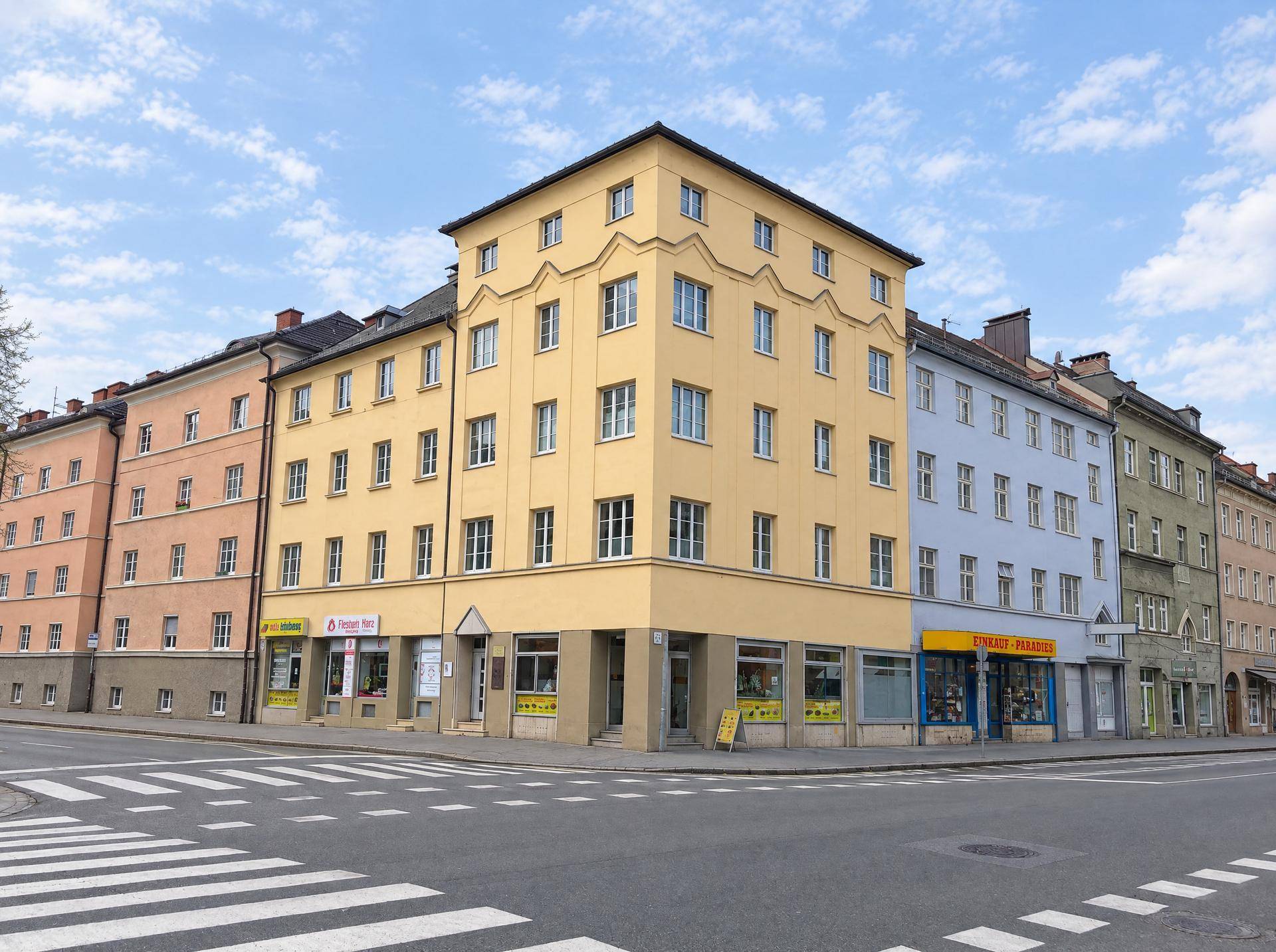 Mehrstöckiges Gebäude an einer Straßenecke mit gelber Fassade und Geschäften im Erdgeschoss.