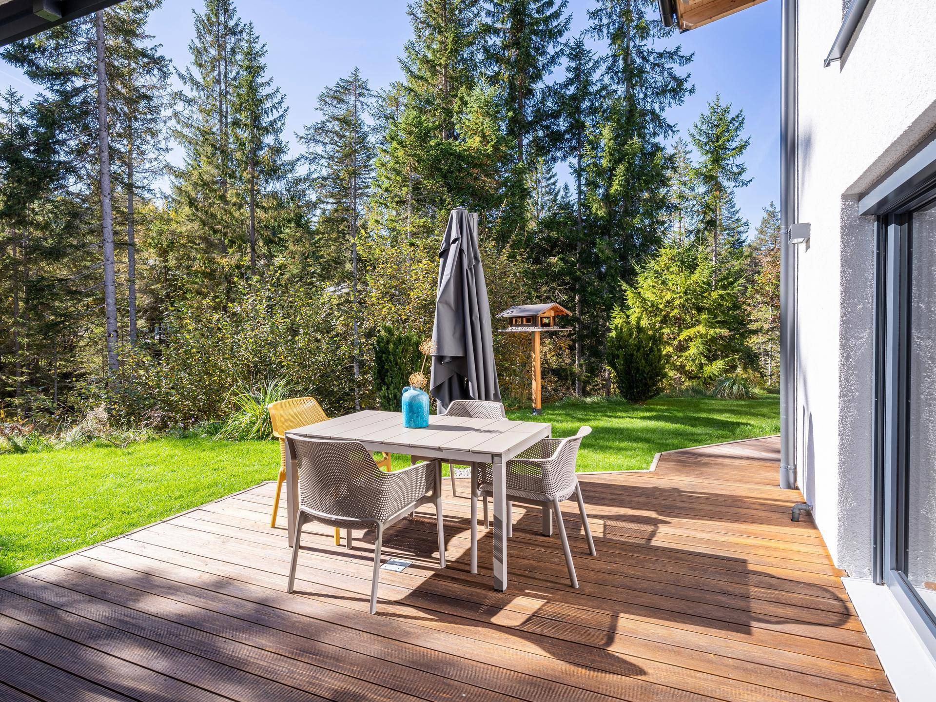 Sonnige Holzterrasse mit Esstisch und Stühlen, umgeben von grüner Natur.