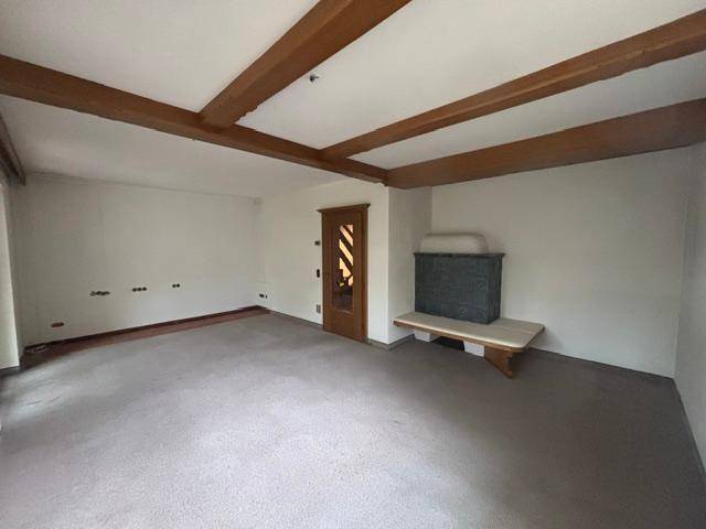 Geräumiges Wohnzimmer mit sichtbaren Holzbalken, Teppichboden und Kachelofen.