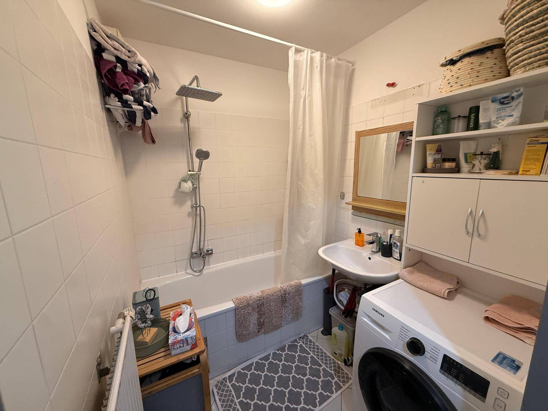 Funktionales Badezimmer mit Badewanne, Duschkopf und Waschmaschine, hell gefliest.