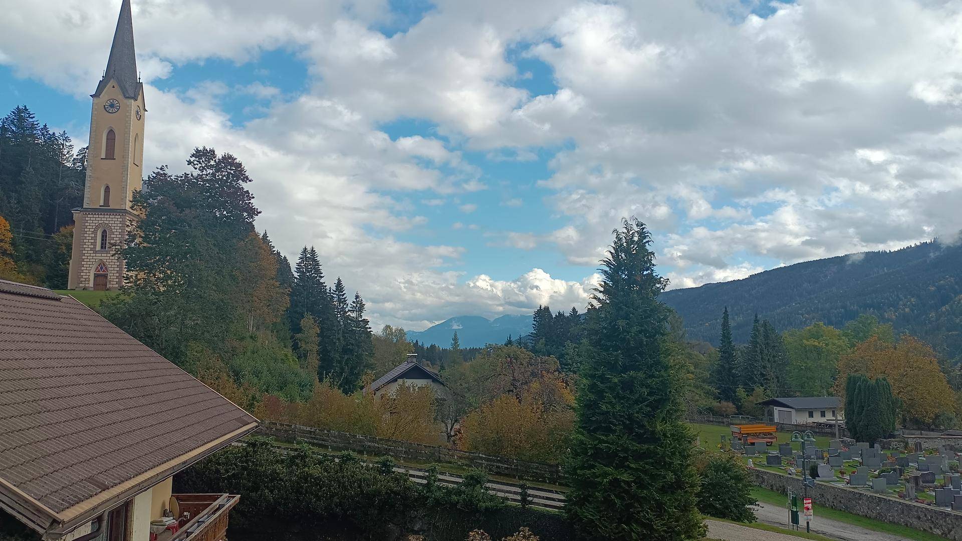 Panoramablick auf ein malerisches Dorf mit Kirchturm, umgeben von Bergen und einem Friedhof.
