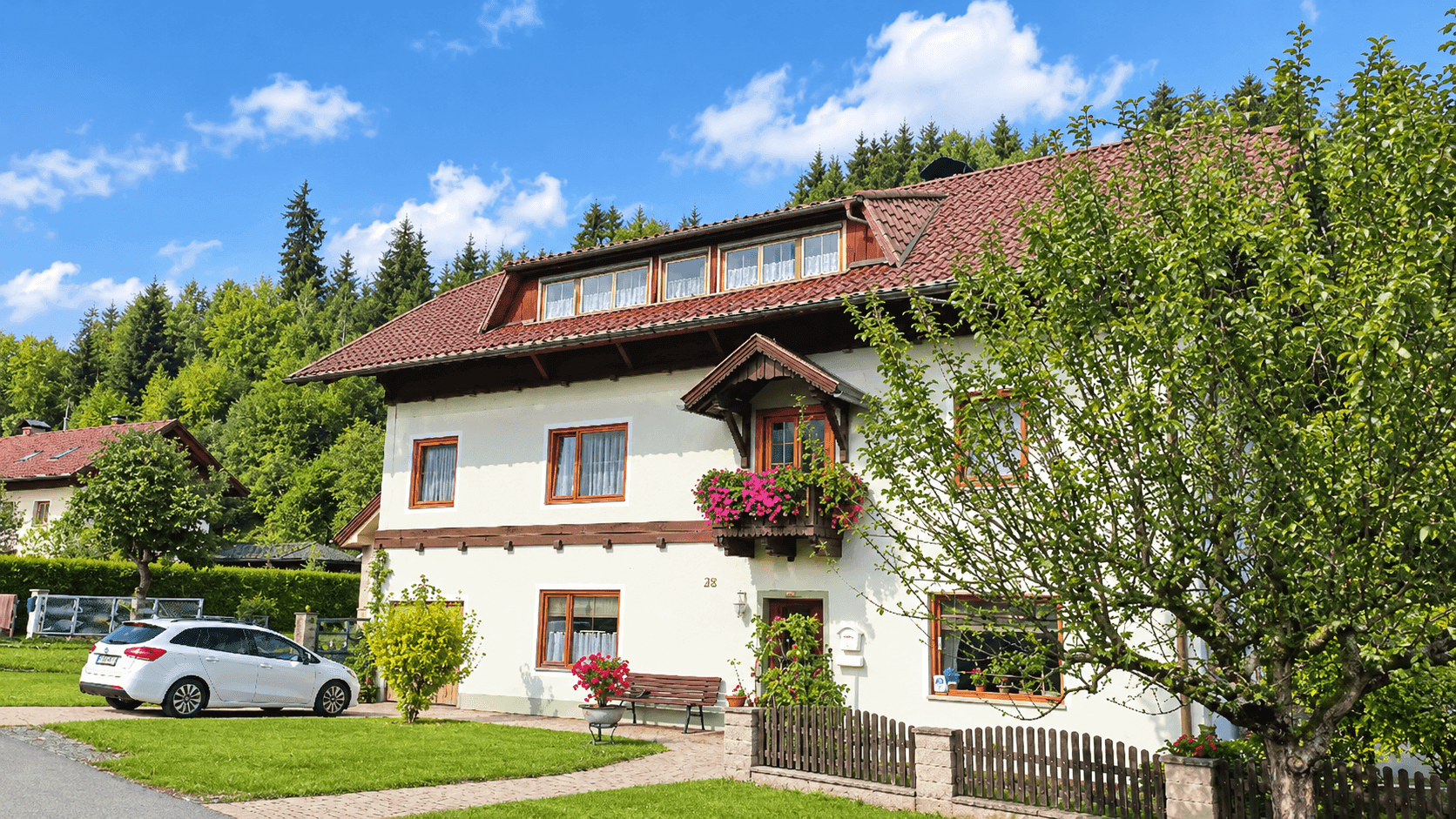 Traditionelles Haus mit Blumenbalkon und gepflegtem Vorgarten unter blauem Himmel.