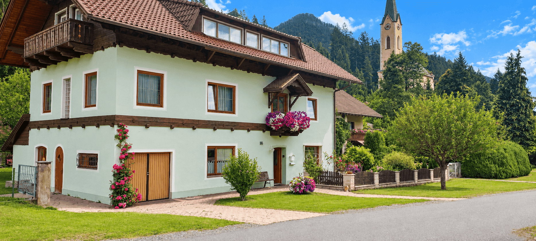 Traditionelles Haus mit hellgrüner Fassade, rotem Ziegeldach und Blick auf die Berge und eine Kirche.