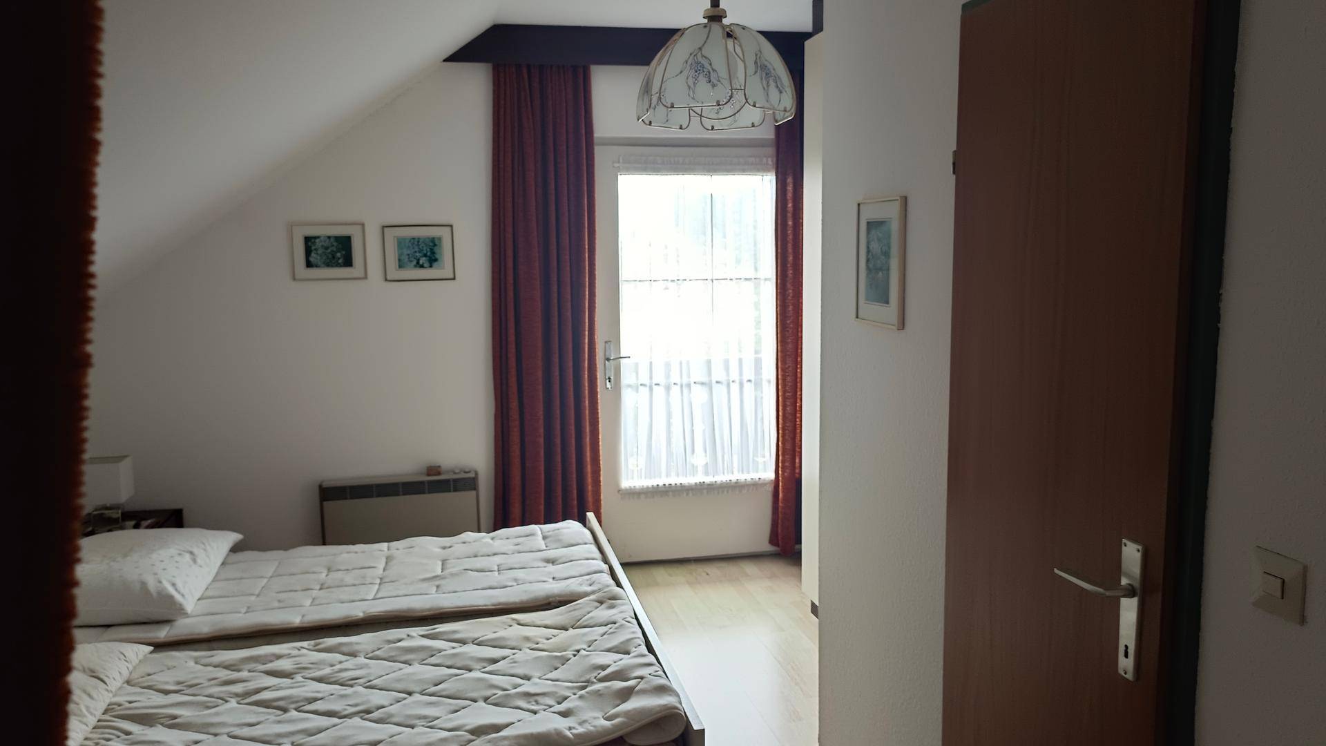Helles Schlafzimmer im Dachgeschoss mit Bett, Heizkörper und Fenster mit Vorhängen.
